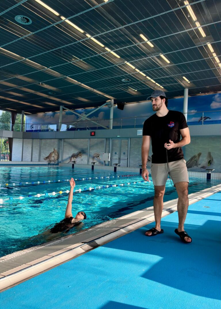 A swimming coach standing poolside in an indoor swimming pool, giving swimming lessons to a swimmer practicing backstroke near the edge of the pool under a well-lit roof.