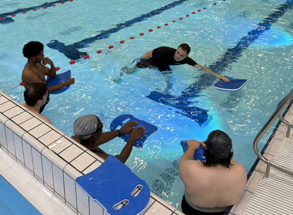 Instructor outlining freestyle mechanics to four adult swimmers using kickboards